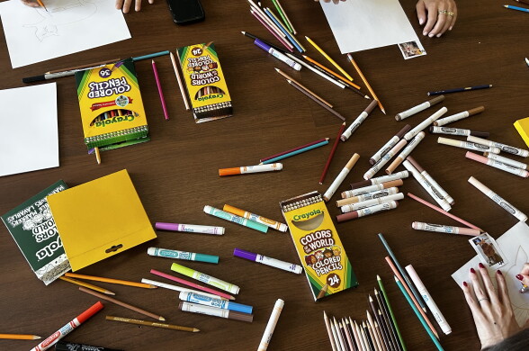 Colored pencils and markers scatter across a wooden table beside open Crayola boxes and sheets of drawing paper.