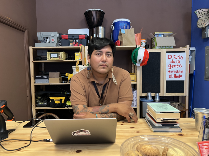 Manager of the lending library César Montero sits behind his check out desk with a rack of items waiting to be checked out from the library behind him.