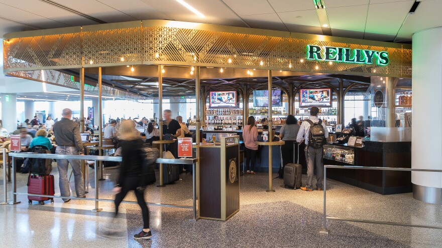 A view of Reilly's Irish Pub in LAX. It's an open restaurant area with a gold lining along the roof. It reads Reilly's in neon green letters as people sit and walk by.