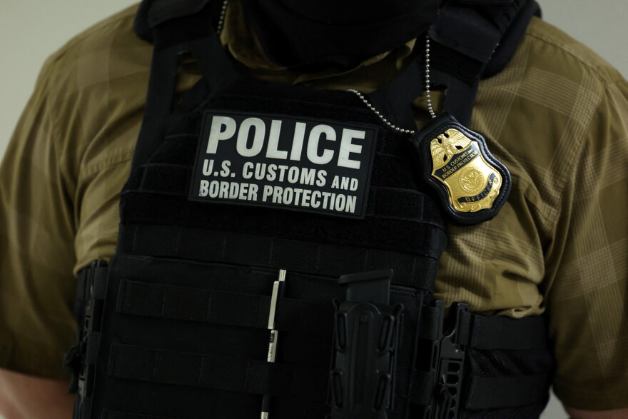 The chest of a person with a black vest that says in large white letters "Police, U.S. Customs and Border Protection." A badge hangs over the vest.