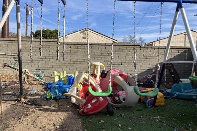 A blue and white swing set with green swings. Half the ground on the left side is covered in sand. The right side is covered in green fake grass. There are three swings on the swing set, but only the middle and right hand one are in tact. The swing on the left has just chains and no swing seat. The chains look charred. Behind the swing set, a children's red plastic truck is semi-melted. A tangle of other plastic colorful toys are behind it. Branches and ash is strewn across the ground.