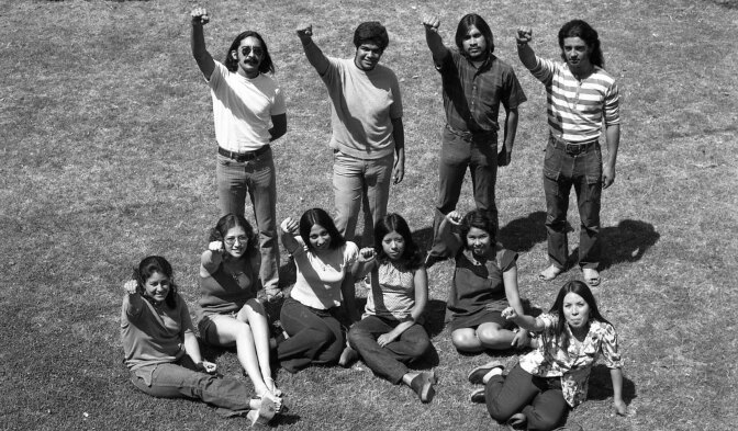 A black and white photo of 10 young adults in 1970s clothes. Their fists are raised.