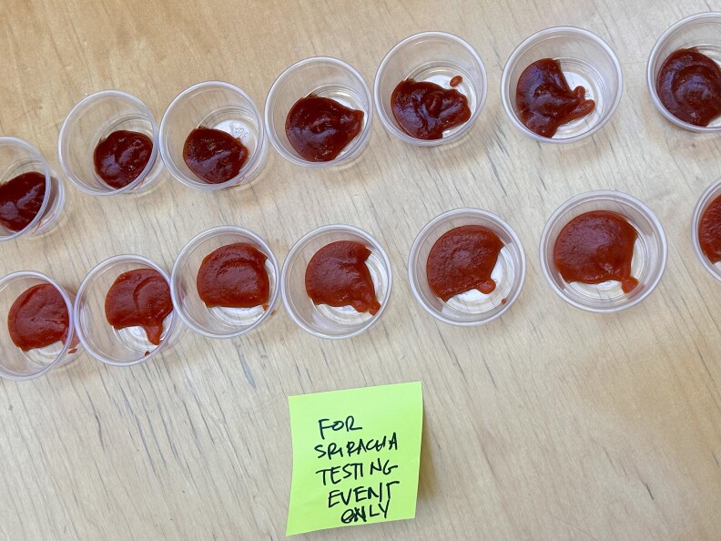 Two lines of clear condiment cups filled with red sauces. The top line of sauce looks darker in color. Underneath all the cups is a bright green sticky note that says "for sriracha testing event only"