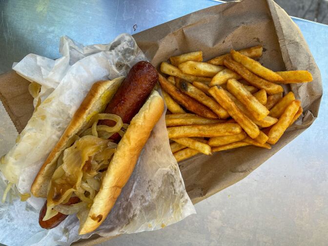 A large dark brown cooked hot dog inside a pale yellow hot dog bun topped with cooked white onion sits over a white to-go paper wrapping containing various grease spots. On the right is a large portion of yellow-gold-colored French fries placed on top of a flattened brown paper bag. 