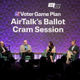 A photograph of a crowded, darkend room: The stage is dramatically lit in purple and there are four people sitting on stage in green club chairs, holding microphones.