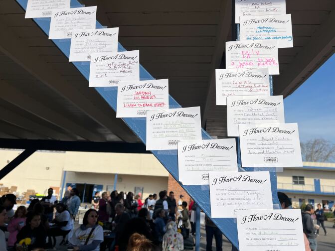 About a dozen certificate-style signs that read I Have A Dream! are taped on a support beam outside a school where dozens of people are gathered on a school playground. Each includes spaces filled in by children that say 'My Name Is,' 'I Am From,' and 'My Dream for the World Is"