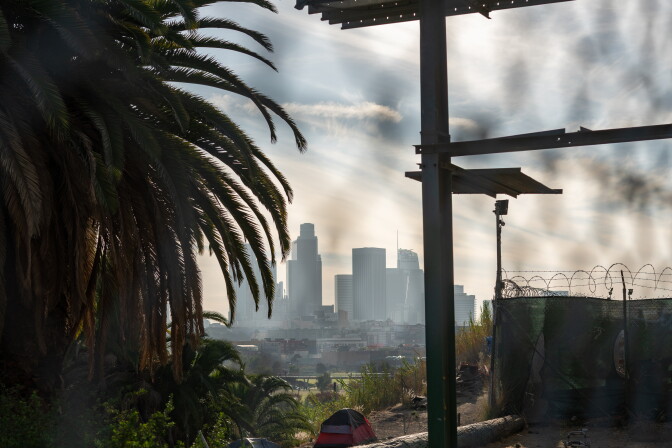 Here's a photo of the downtown Los Angeles skyline along with taken from the view of the North Broadway bridge. There is a palm tree, tents and a billboard in the foreground. 