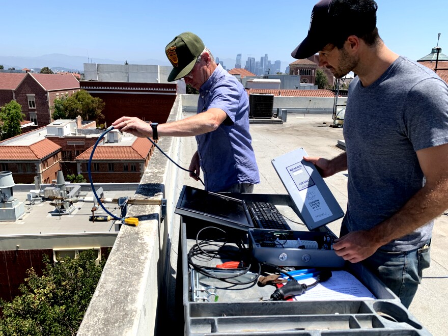 An image of USC professor William Berelson, wearing a green baseball cap and holding a cable, stands beside his  research assistant, Nick Rollins, on the rooftop of a school in Los Angeles. Rollins stands in front of a laptop and holds equipment to attach an air pollution sensor to the side of the building. 