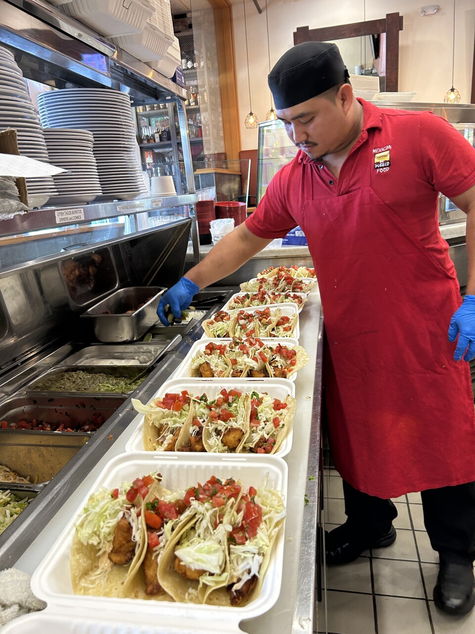 A man in a red apron and a black chef's hat grabs for a handful of cut limes as he attends to tray after tray of fish tacos that are lined up in front of him, ready to go for hungry customers. 