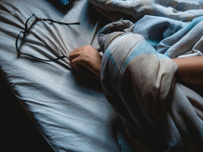 An arm draped in a sheet rests on a bed close to a pair of glasses.