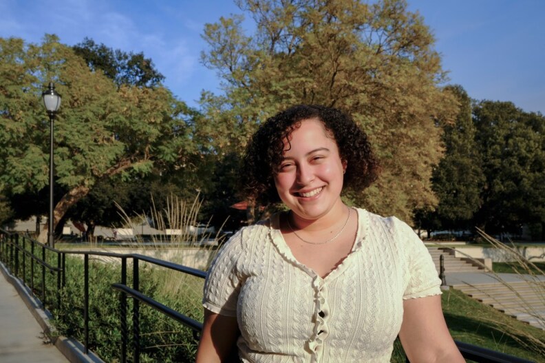 A young woman with pale skin and short brown curly hair smiles at the camera. She stands on a walkway in front of green trees and bushes. She wears a pale yellow ruffled, short-sleeved shirt.
