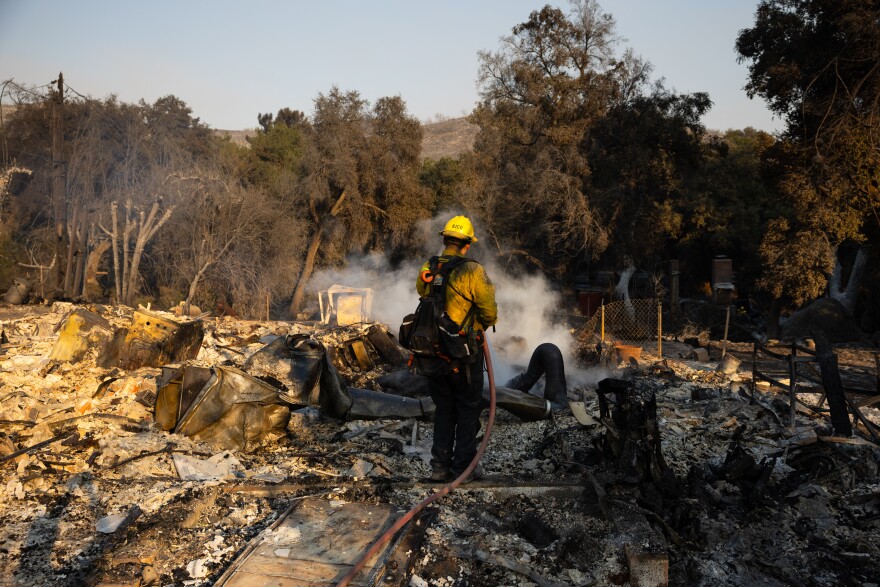 A firefighter stands with his back to the camera in a field with smoking debris.