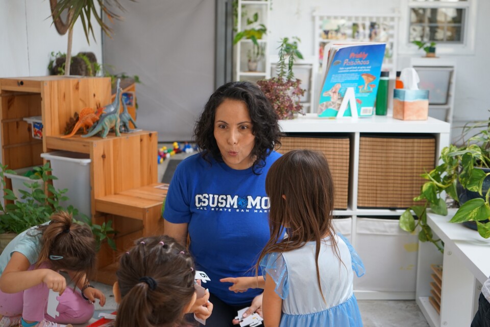 A woman with a medium skin tone and short curly brown hair makes a smiley face at a young student with brown hair. Two other young children are in the frame, their faces out of view. Blocks, baskets and a children's book are behind the woman, as are toy dinosaurs. To her left, a green plant sits on a counter. Her shirt reads "CSUSM MOM."