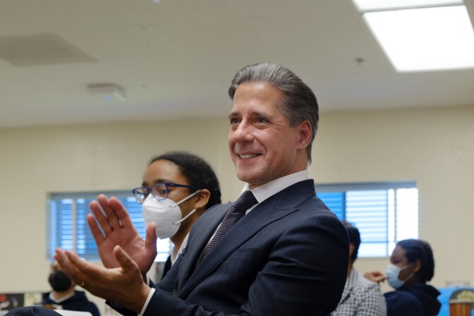 Alberto Carvalho claps wile sitting in a room wearing a dark suit and tie.