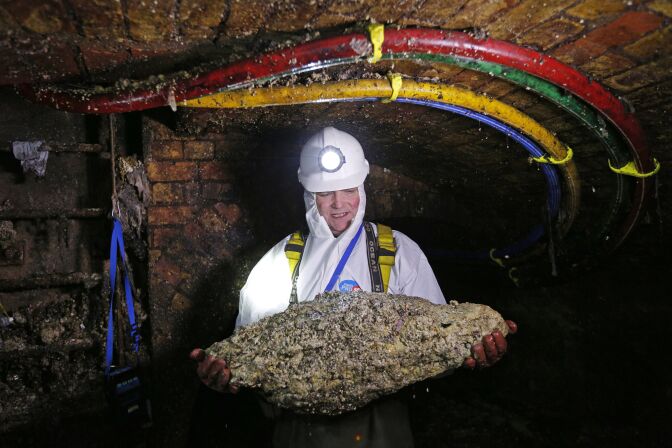 A man in a white full-body suit, headlamp and gloves stands in the middle of a sewer holding a pale conglomerate that's about the size of a large toddler.  