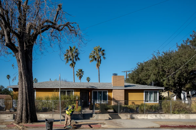 A person wearing a yellow safety vest rakes debris off the curb of a street in front of a yellow house and tree.