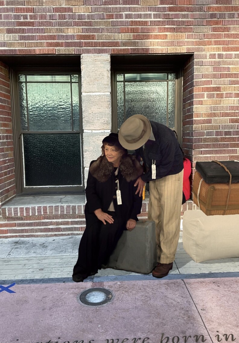 A screenshot from an augmented reality app shows an older woman in a fur coat sitting on a suitcase while a man standing next to her provides comfort.
