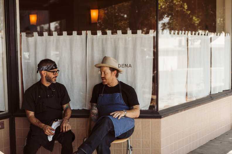 Two chefs in aprons sit outside a storefront, mid-conversation, one in a wide-brim hat and the other in a bandana, the afternoon light bouncing off the café’s tiled exterior.