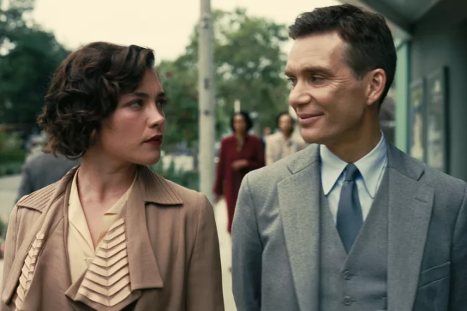 Florence Pugh and Cillian Murphy walk down a street together. Their clothes and surroundings indicate that they are in the 1950s. Pugh wears a tan collared coat with a champagne blouse underneath. She looks over a Murphy with sternly, seemingly annoyed at him. Murphy smiles back at her through closed lips, seemingly amused at Pugh's disdain. He wears a grey three piece suit and tie. 