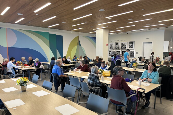 A senior center room filled with at least 7 dining tables with 8 seats each. Older adults are sitting across the tables, some with lunch trays and paper cups in front of them. At least two men can be seen walking the room in aprons and hairnets, handing out food to the older adults.