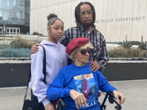 Takar Smith's family comes together for a photo outside LAPD headquarters on Friday, January 13. Smith's niece, Rashi wears a purple hoodie. His brother, Raischard, wears glasses and a plaid shirt. Raischard has his hand on their mother, Alicia Smith, who wears a blue shirt that has an image of Takar on it. 
