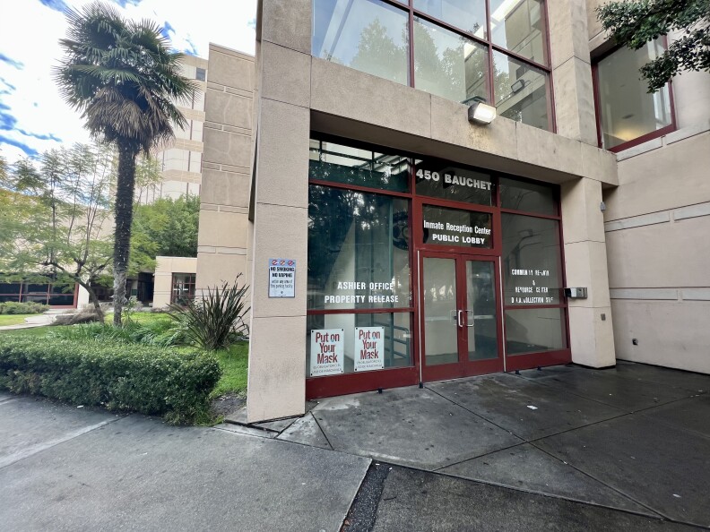 We're looking at the entrance to the downtown jail public lobby and inmate reception center. A concrete beige building faces us, with glass windows framed with red. The door has white signs like "put on your mask". The building is part of a large complex -- we can see other beige buildings beyond the reception center. A palm tree is to the right of the building, and there are green grass and shrubs.