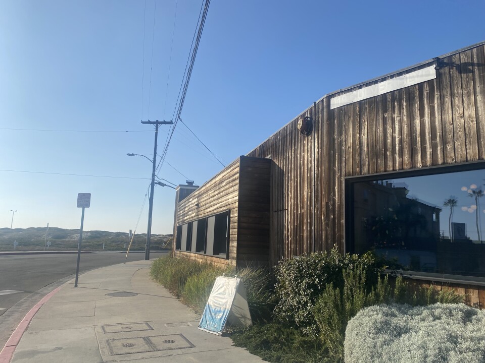 The wooden-clad exterior of a restaurant that is sun-drenched, as it looks out toward the beach. 