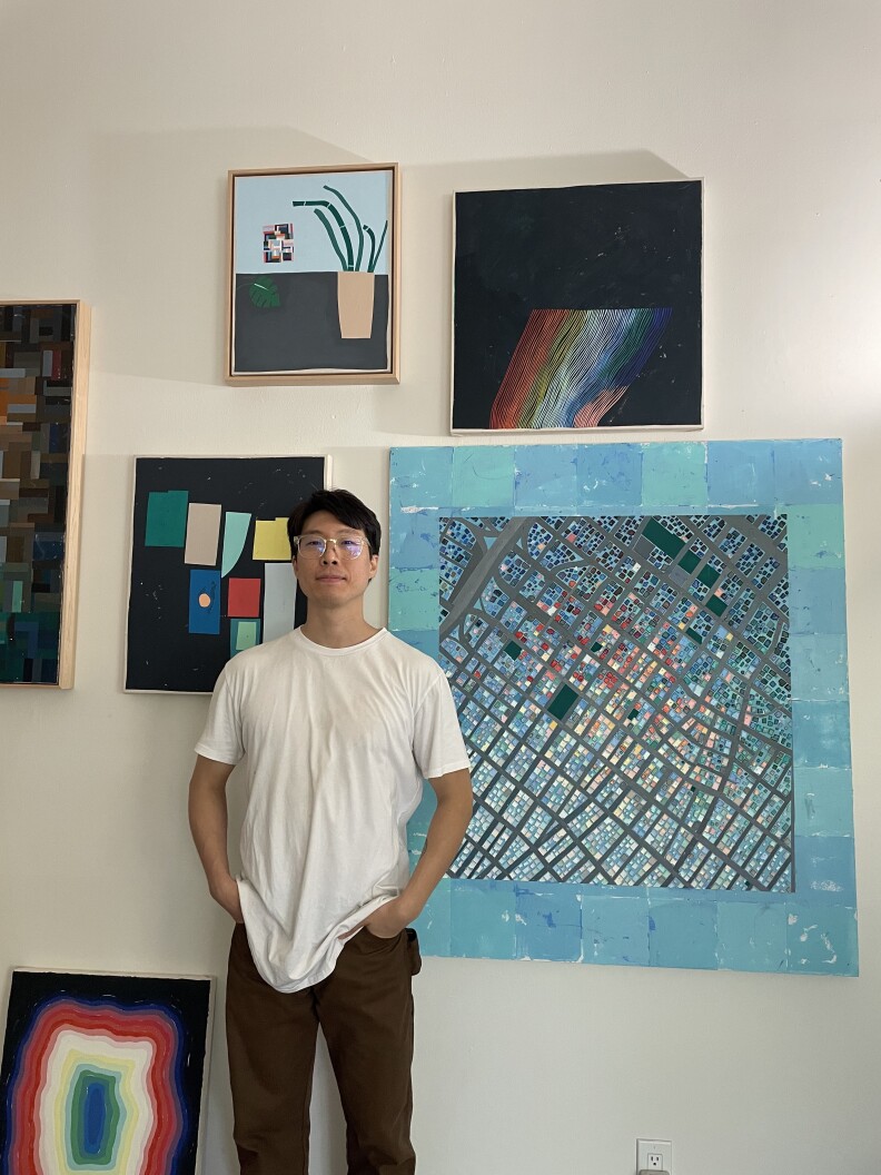 A man with light skin tone stands in front of some of his art hanging on a white wall.