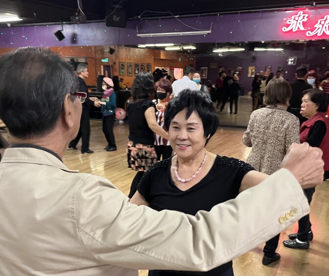 An Asian woman with short black hair dances on a busy dance floor with an Asian man in a cream blazer with his back to the camera. 