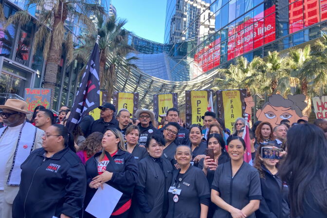 A crowd of people are standing before a large hotel in downtown Los Angeles. There are several signs and a flag as  well as palm trees lined behind them with a blue sky peaking behind them. The front row shows five women smiling. 