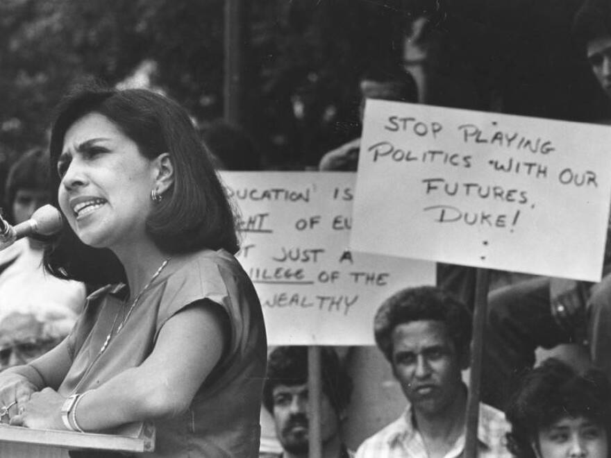 A black and white photo of a young Latina woman with short dark hair wearing a medium dark short sleeve dress. She stands at a podium speaking into a microphone with a serious expression. Behind her there are Latino people holding signs that read "Stop playing politics with our futures Duke!" and "Education is a right for everyone, not just a privilege of the wealthy."