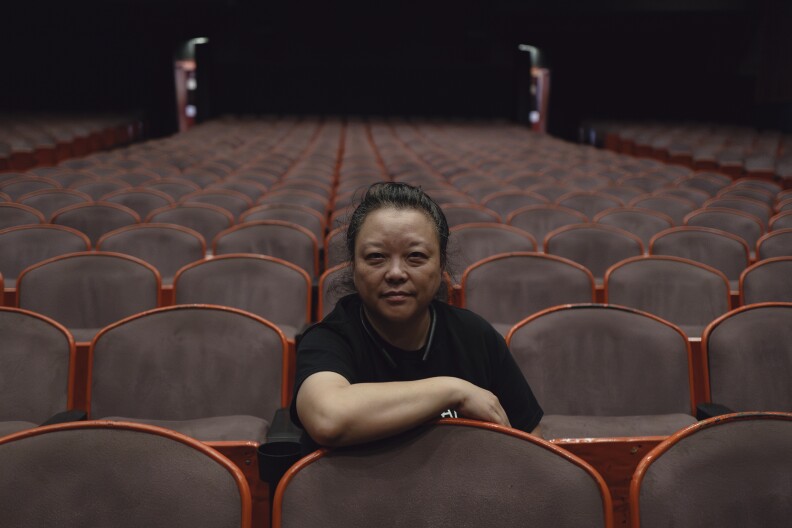 An Asian woman with her hair pulled back and wearing a black t-shirt sits alone in a theater auditorium with hundreds of empty seats. She leans forward with a slight smile and rests her arm on the seat in front of her.