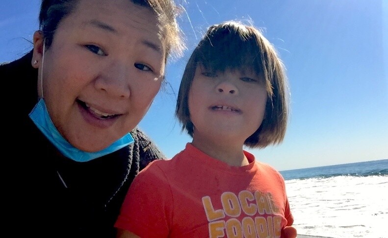 An Asian American woman with a blue face mask pulled down around her neck stands beside her daughter, who has a pageboy style haircut and is wearing a red t-shirt, on a beach.
