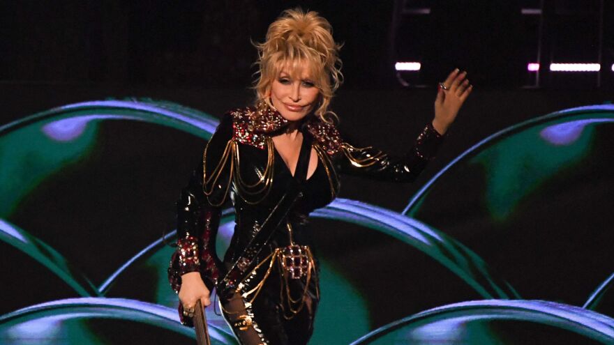 Dolly Parton, a white woman in her 70s, stands on a stage and waves to the audience. She appears to hold a microphone in her other hand. She wears a tight-fitting dark gold dress, and her blonde hair is in a messy updo.