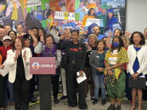 A group of people stand behind a podium with a sign that reads "CHIRLA." They also stand in front of a painted mural. Some people are raising a fist upend others are clapping. 