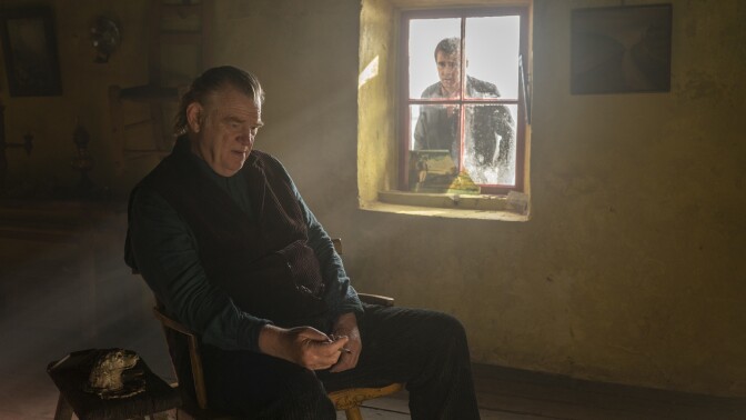 Brendan Gleeson sits in the middle of a room on a wooden chair next to a small end table with a large window to his right. The room is bare with nothing on the walls except a small landscape painting next to the window on its right side. The window provides the only light, the rest of the room and its limited possessions fading into shadow. Gleeson stares at the floor, his lips slightly parted. His hands rest in his open lap as he loosely holds a pen. He is wearing a dark blue long sleeve with a brown buttoned sweater vest and black pants. Gleeson's absent expression shows that he is unaware or unconcerned by Colin Farrell peering in at him from the large window. Farrell stares at Gleeson from outside the room his expression confused, the light of the day shining behind him. 