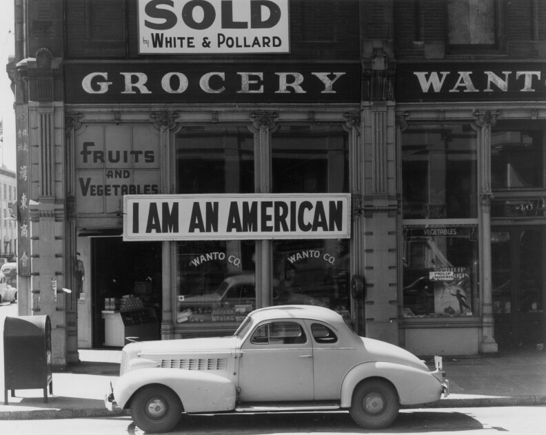 A black and white photo from 1941 shows a storefront with a car parked in front. A large sign hung on the building reads "I am an American"