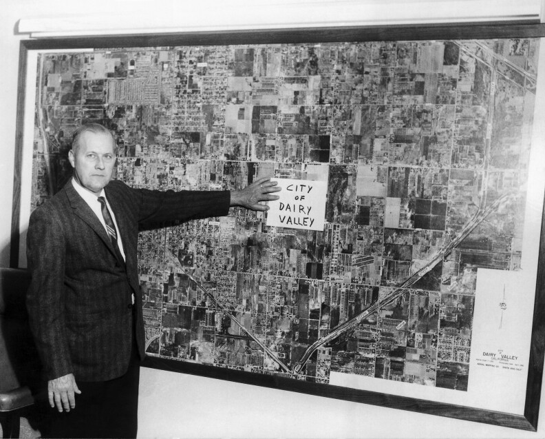 A black and white archival look at a white man in a suit standing next to a large aerial image that shows a wide area of undeveloped land with note on it that says city of dairy valley.