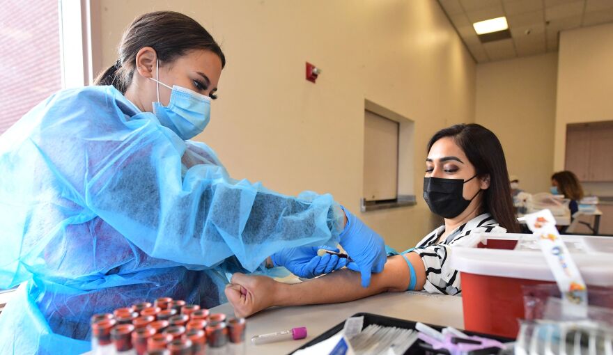 A female phlebotomist dressed in blue PPE draws blood from a woman's arm inside a clinic.