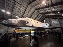 A massive white and black retired space shuttle towers over a crowd of museum-goers. 