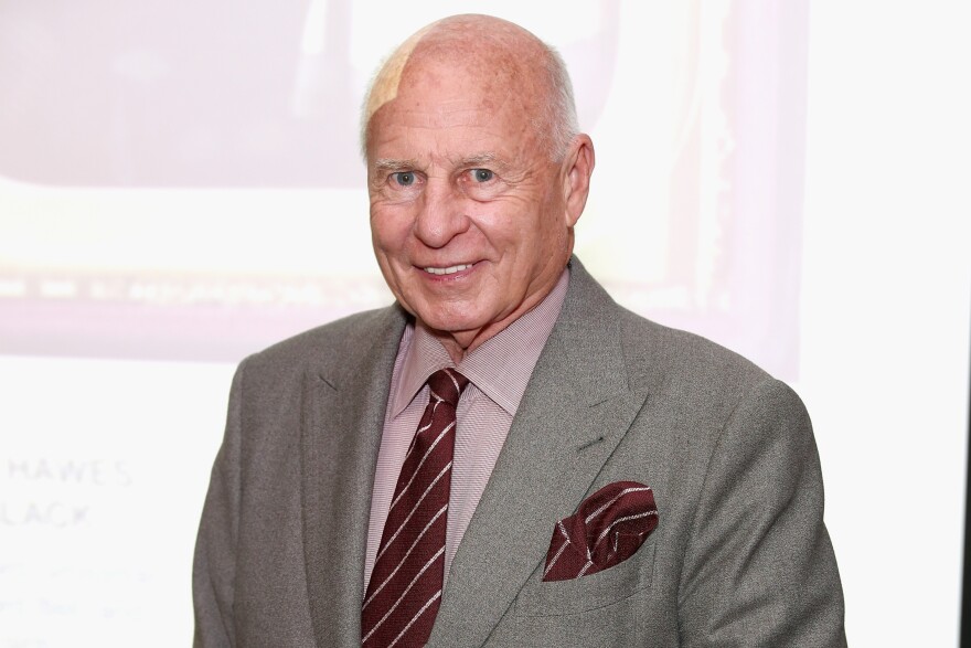 A photo of an older white man giving a slight smile and looking at the camera. He appears to have a bandage on his head. He is wearing a gray suit with a maroon striped tie and pocket square.