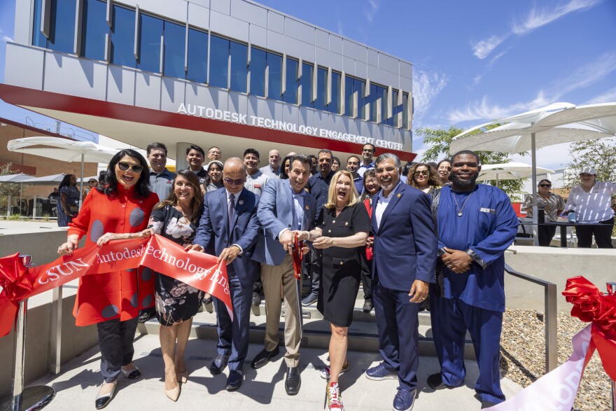 A large group of people gather in front of a gray building with red trim. The building displays the name "Autodesk Technology Engagement Center." A red ribbon in front of the crowd is cut in half.