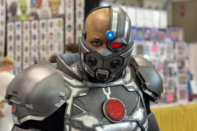 A brown-skinned man in a Cyborg cosplay outfit, with a robotic covering over his mouth (and a COVID-19 mask peeking through). He stands in front of a brightly colored convention booth in soft focus in the background.