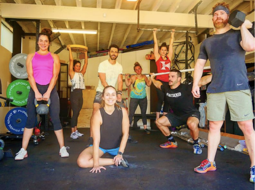 Friends on the workout bubble pose for a picture in a garage with workout equipment, including barbells and free weights.