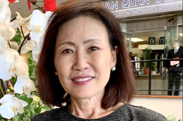 An Asian American woman in a checkered sheath smiles at the camera. In the backdrop are a vase of flowers and a tailor shop that is part of a shopping mall.