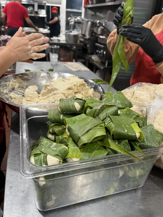 Two containers sit on a metal table. One is a large bowl full of masa, the other contains wrapped balls of masa with the green corn leaf. Two hands hover over each.
