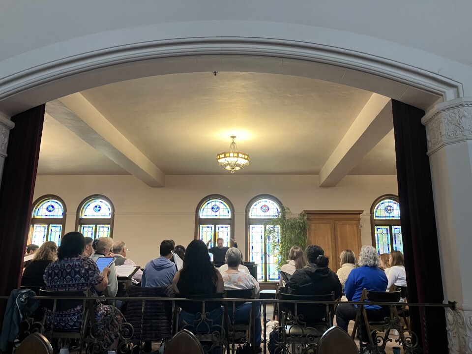 A light-skinned man stands in front of a group, hands raised above his head. He stands in front of two stained windows, with an indoor plant. Rows of people sit in chairs looking at him.
