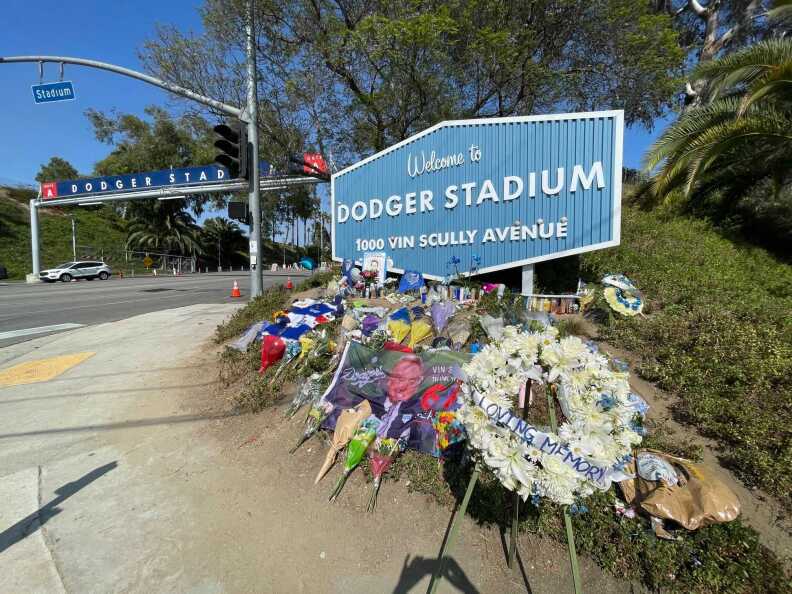 A memorial of candles, flowers, signs and more set up in front of a welcome sign at Dodger Stadium, which includes the address 1000 Vin Scully Avenue.