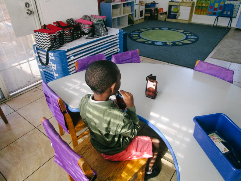 In this image, a child sits at a table in a classroom while facetiming another child on a smart phone.