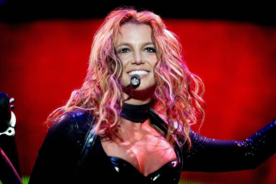 Britney Spears performs in a dark outfit, a red backdrop is in the back.